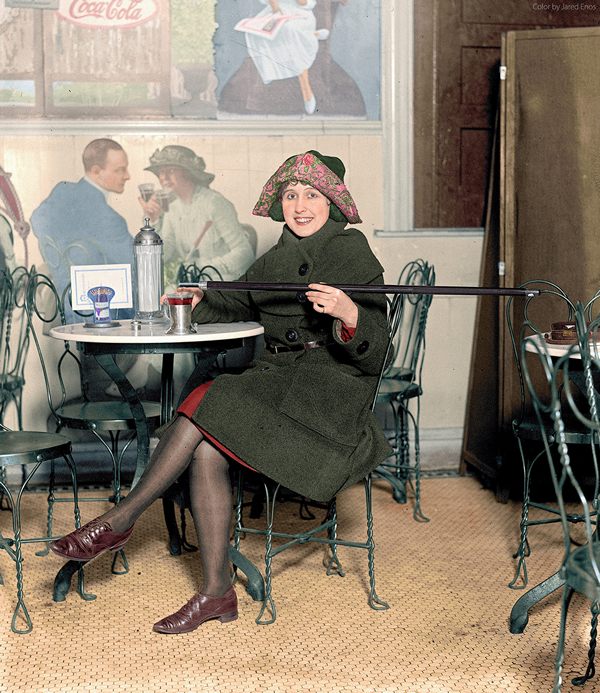 Woman seated at a soda fountain table is pouring alcohol into a cup from a cane, during Prohibition; with a large Coca-Cola advertisement on the wall